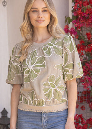 Woman wearing a beige blouse with green floral patterns standing in front of red flowers.